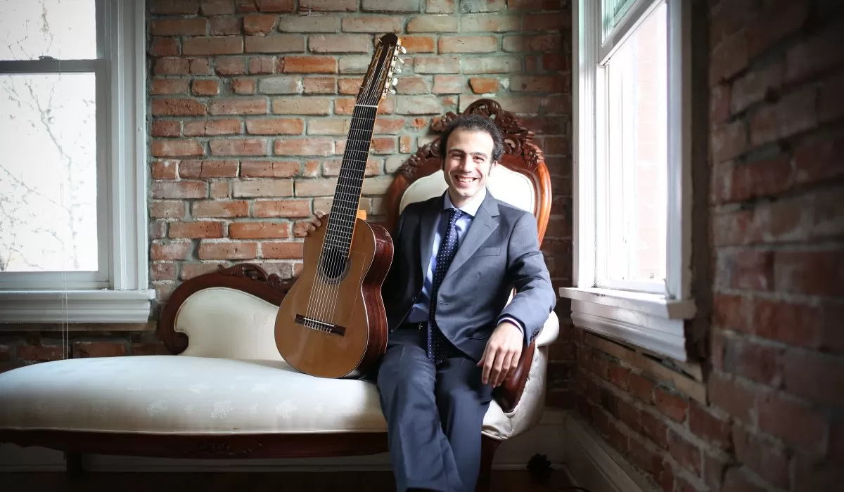 A smiling musician in a suit seated beside a classical guitar in a brick-walled room.