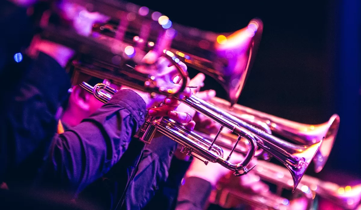 Close-up of brass jazz instruments being played under vibrant purple and pink stage lighting.