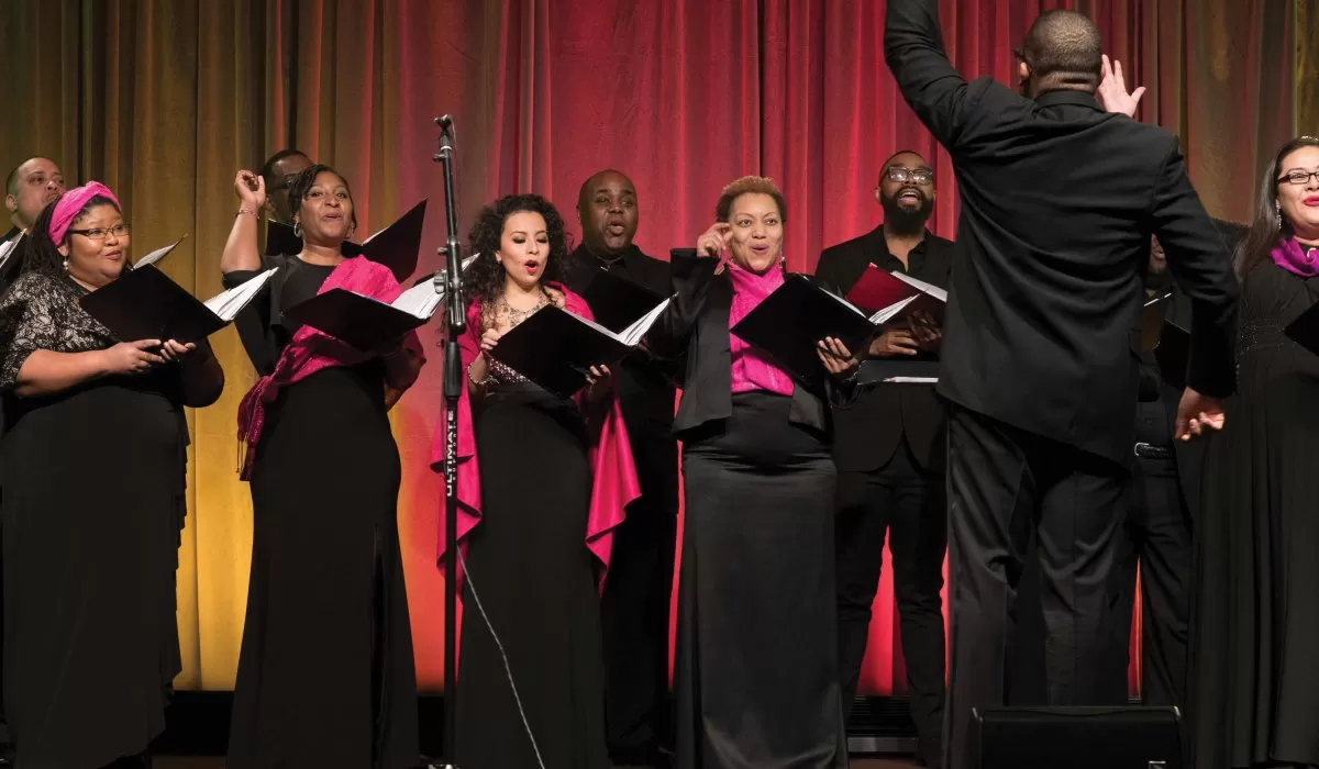 Choir members singing onstage with a conductor in front of red curtains.