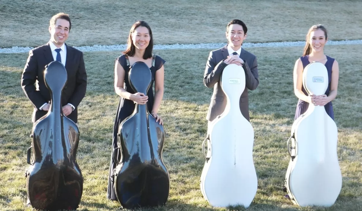 Four musicians stand outdoors on a grassy lawn, each holding a cello case.