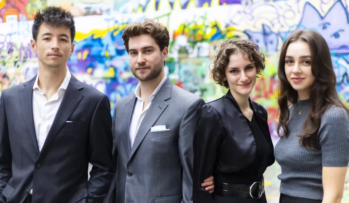 Four musicians pose for a group portrait in front of a colorful graffiti-covered wall, dressed in formal and semi-formal attire.