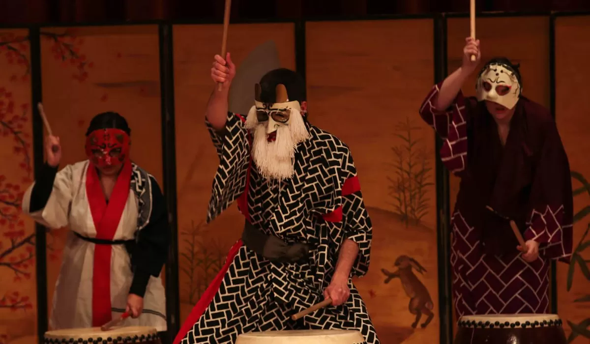 Three masked performers in traditional Japanese-style robes play taiko drums on stage, raising drumsticks in a performance.
