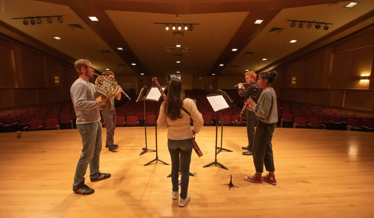Five musicians rehearse on stage in an auditorium, standing in a semicircle with music stands and instruments.