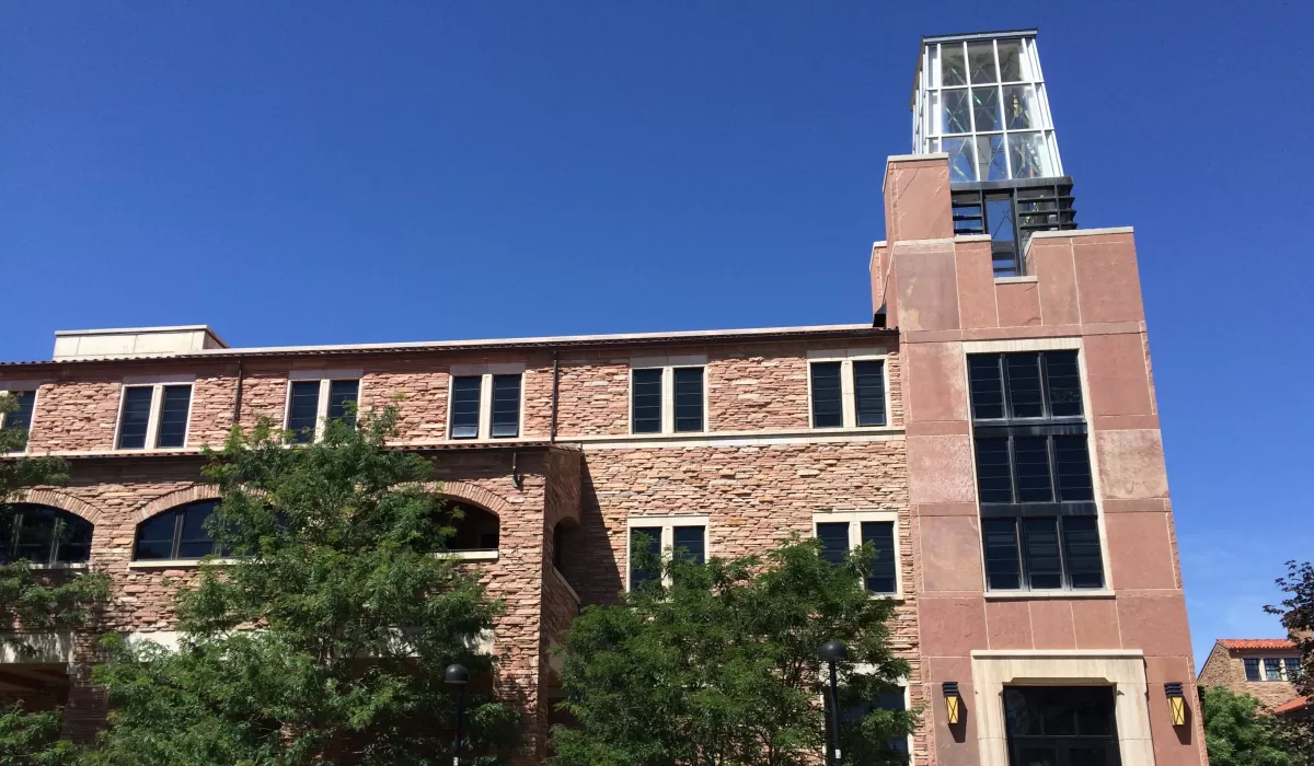 Exterior view of the Atlas building on a sunny day, featuring a stone façade, glass clock tower, trees lining the entrance, and bicycles parked along the sidewalk.