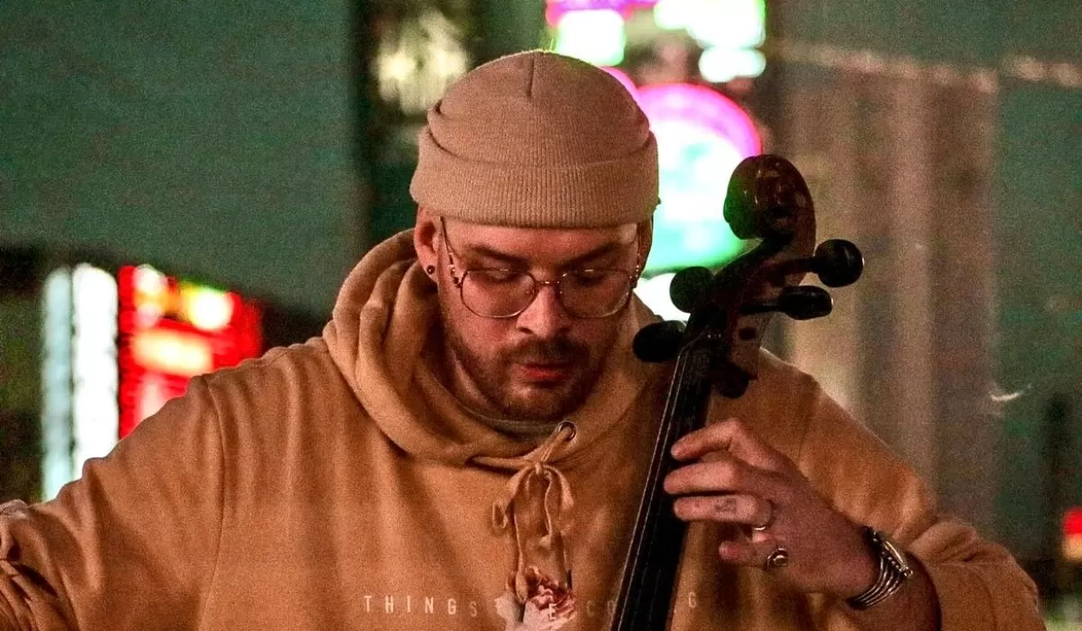 Cellist wearing a tan beanie, glasses and a brown hoodie plays the cello in a nighttime setting with colorful lights in the background.