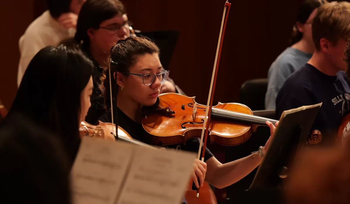 A violinist rehearses with an orchestra, focused on sheet music during a performance.