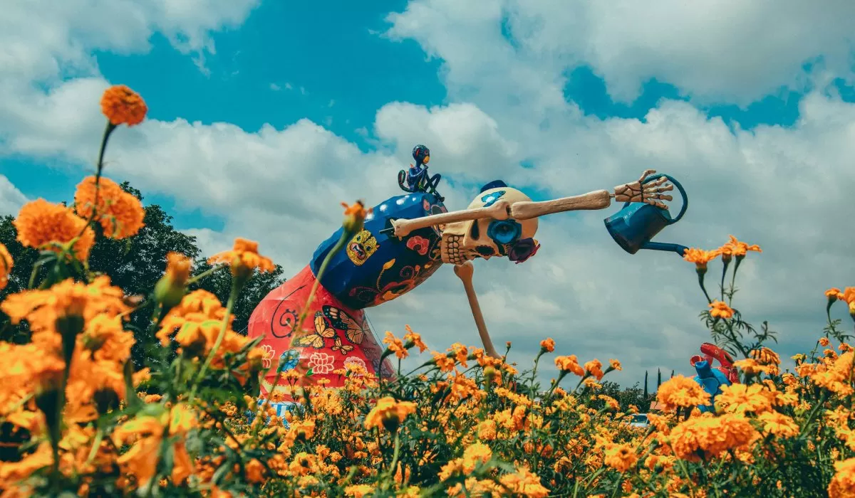 A colorful skeleton sculpture in a field of orange marigolds, watering flowers under a blue, cloudy sky.