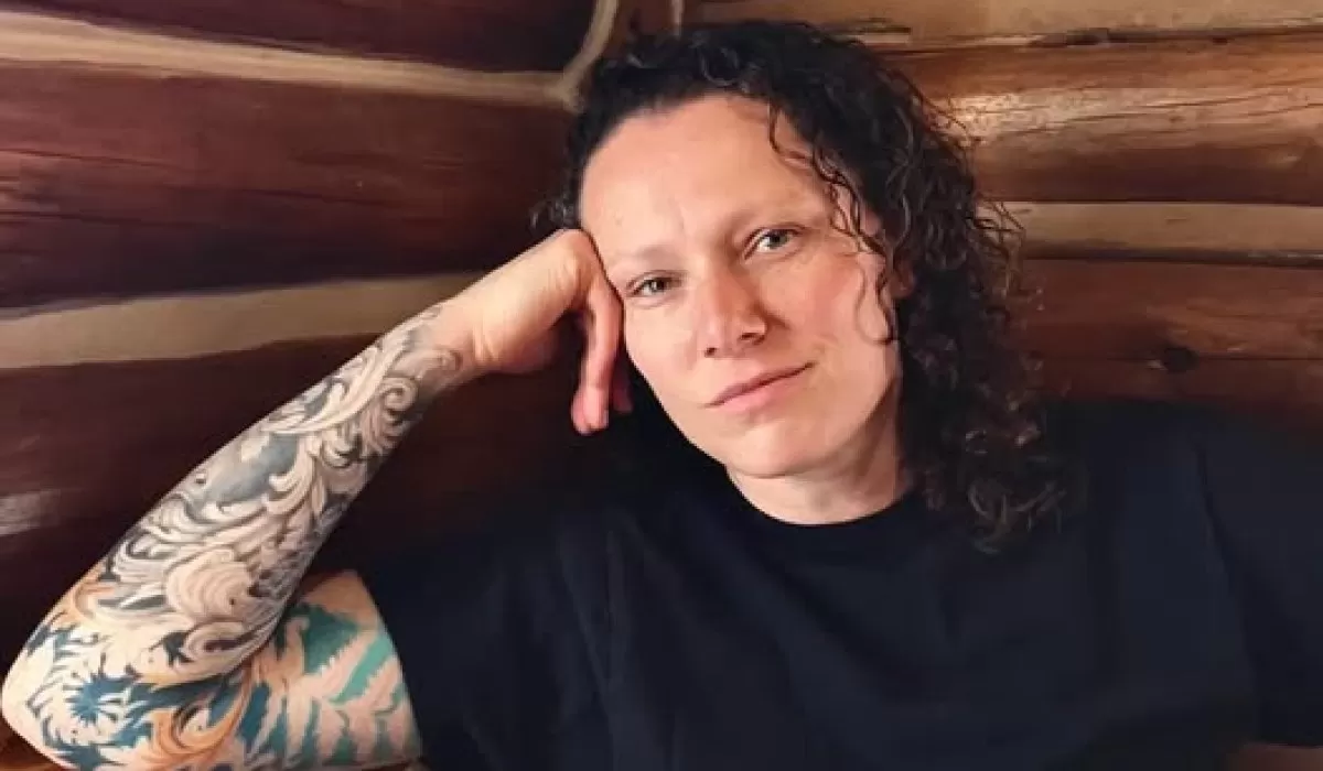 Person with curly hair and a tattooed arm, sitting against a wooden wall.