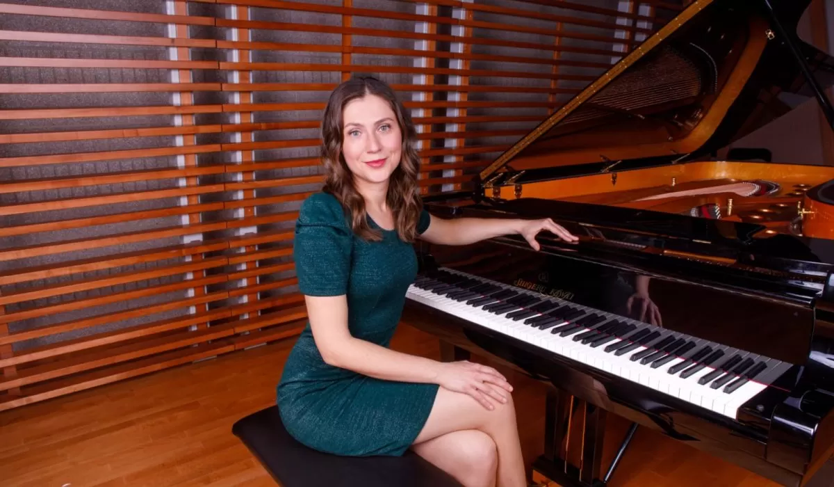 Woman in a teal dress sitting beside a grand piano.