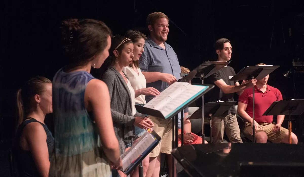Students rehearse a vocal performance on a theater stage, standing behind music stands and singing during a practice session.