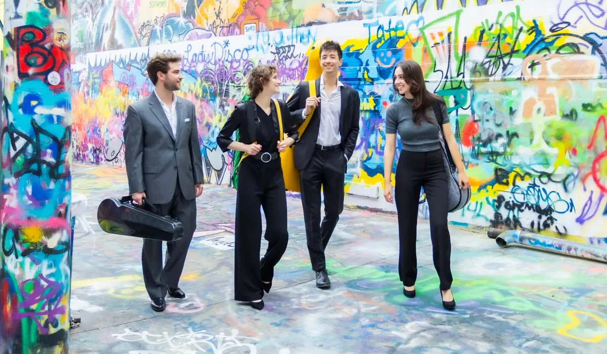 Four musicians walk through a colorful graffiti-covered alley, carrying instrument cases and bags.