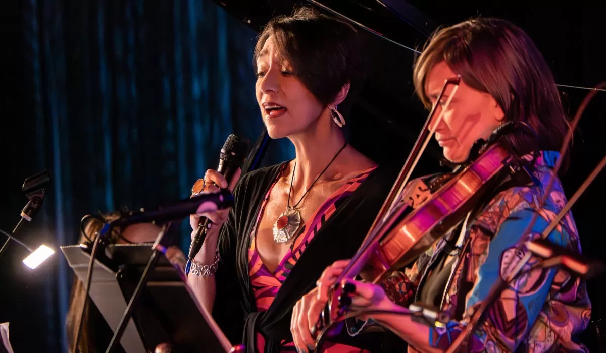Two musicians perform on stage, with a vocalist singing into a microphone beside a violinist, illuminated by warm stage lighting with a grand piano in the background.