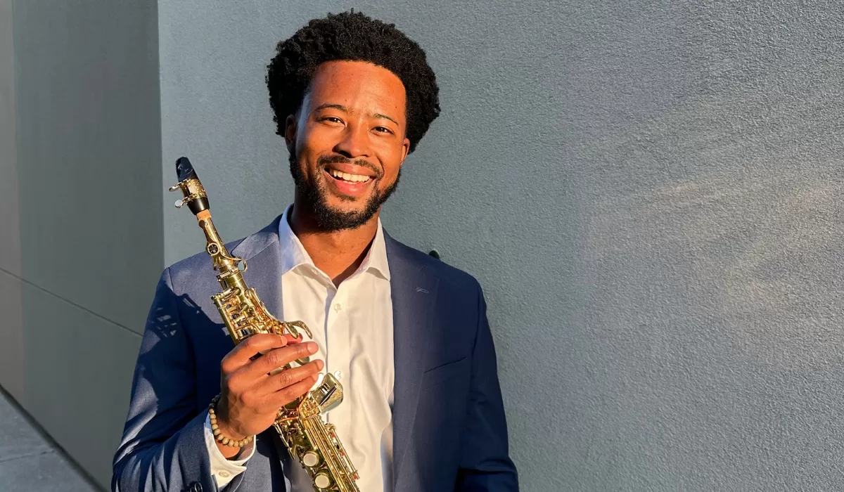 Smiling musician in a suit holding a gold saxophone outdoors against a gray wall.