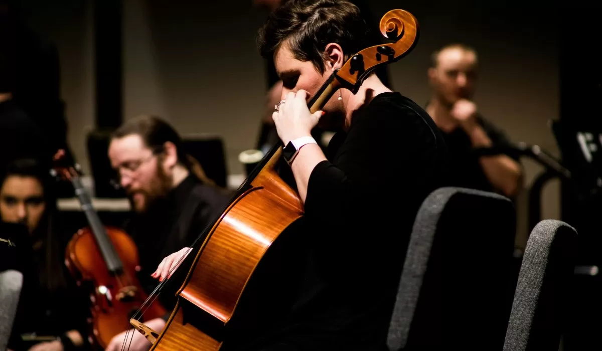 A cellist plays during a performance, with other musicians visible in the background.