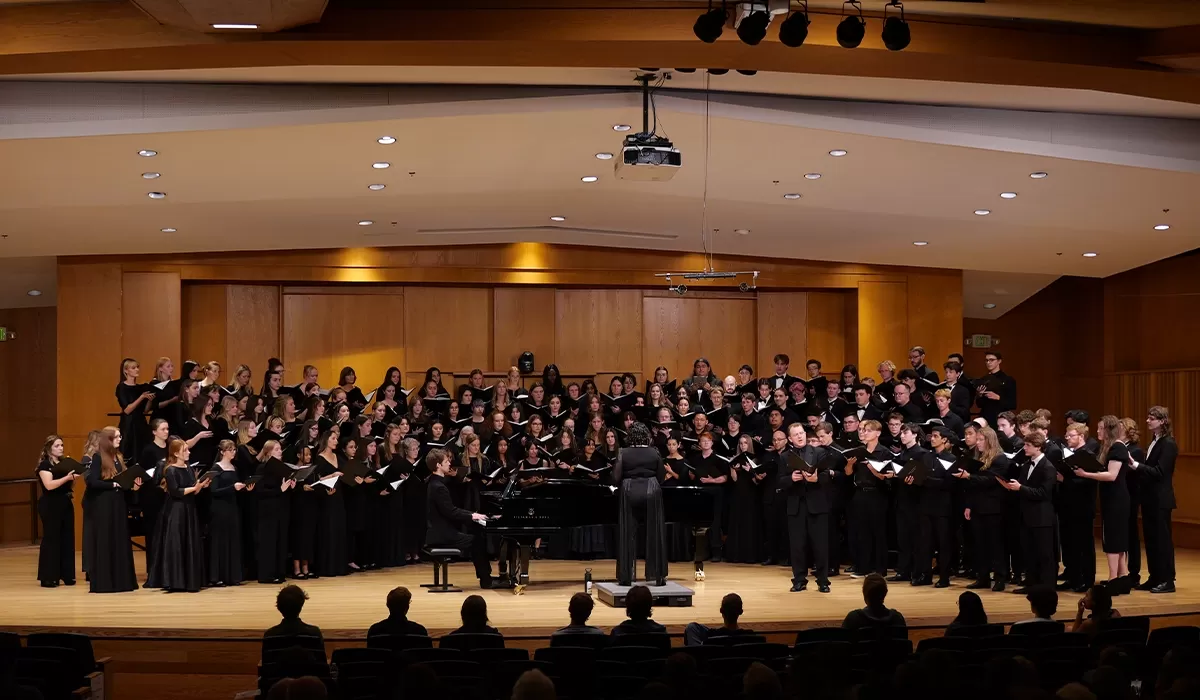 Large choir performing on stage in a concert hall, with a pianist and conductor.