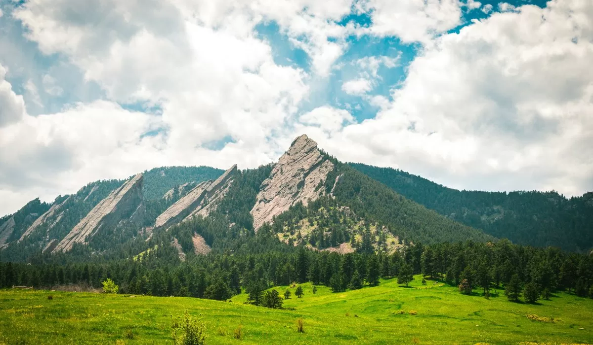 Scenic view of the Flatirons above a green meadow, with pine trees and a partly cloudy blue sky.