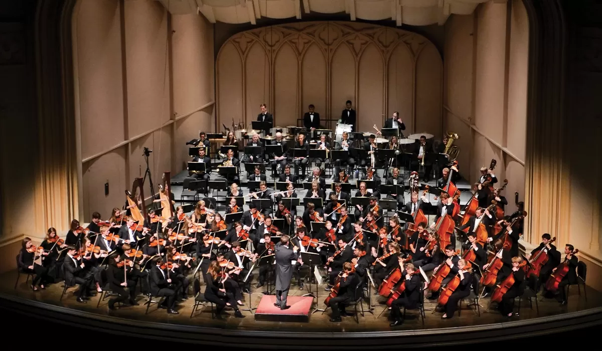 A full orchestra performs on stage under the direction of a conductor, with string, woodwind, brass, and percussion sections.