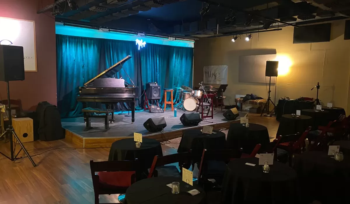 Jazz club interior with a grand piano, drum kit, stage, and tables set for guests.