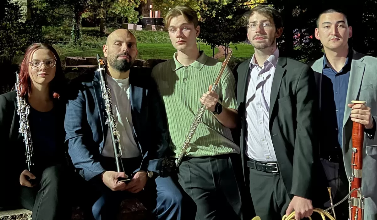 Five musicians pose outdoors at night holding woodwind instruments, including oboes, a flute, and a bassoon, with trees in the background.