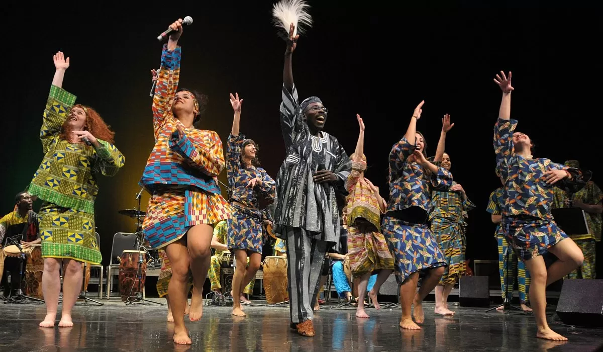Performers in colorful West African-inspired attire sing and dance on stage during a lively musical performance.