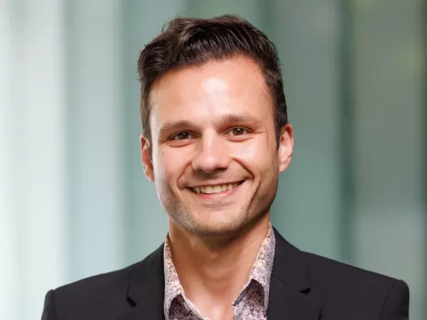 Portrait of a smiling man with short dark hair, wearing a dark jacket and patterned shirt.