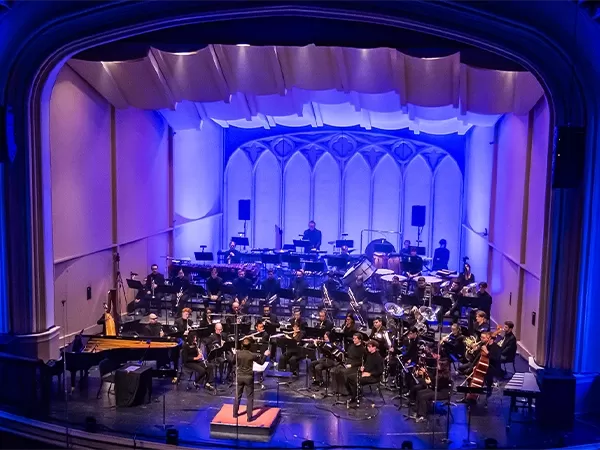 Band orchestra performing on the stage of Macky Auditorium, with a conductor leading musicians