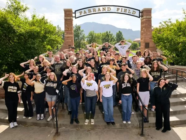 A group of CU Boulder College of Music students pose in front of the Farrand Field entrance, wearing black and gold Buffaloes apparel.