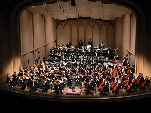 A full orchestra performs on stage under the direction of a conductor, with string, woodwind, brass, and percussion sections.