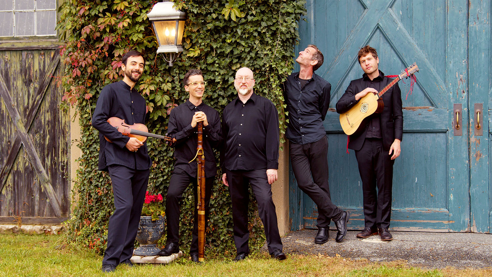 Five musicians with early music instruments standing outside by a barn door and ivy covered wall.
