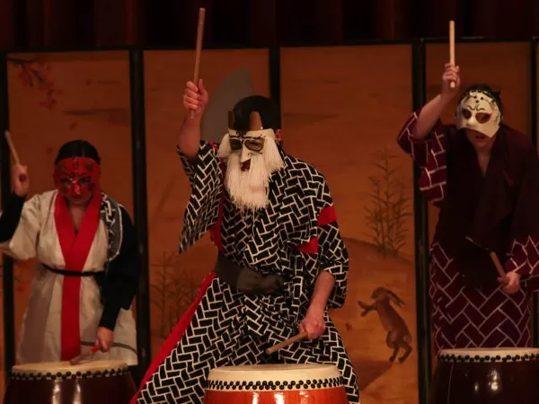 Three masked performers in traditional Japanese-style robes play taiko drums on stage, raising drumsticks in a performance.