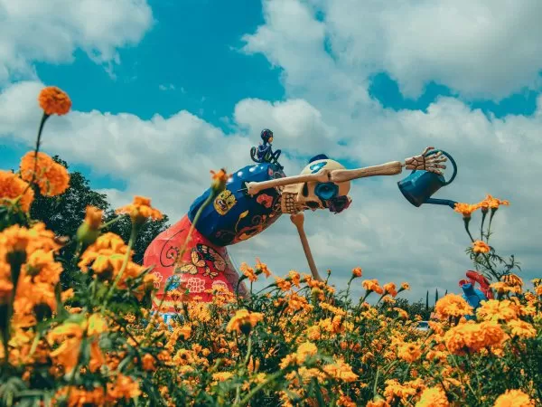 A colorful skeleton sculpture in a field of orange marigolds, watering flowers under a blue, cloudy sky.