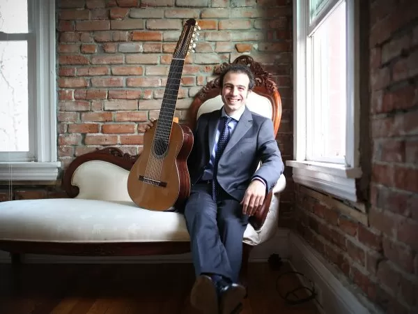 A smiling musician in a suit seated beside a classical guitar in a brick-walled room.