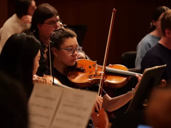 A full orchestra performs on stage under the direction of a conductor, with string, woodwind, brass, and percussion sections.