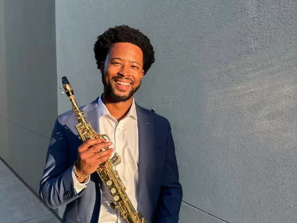 Smiling musician in a suit holding a gold saxophone outdoors against a gray wall.