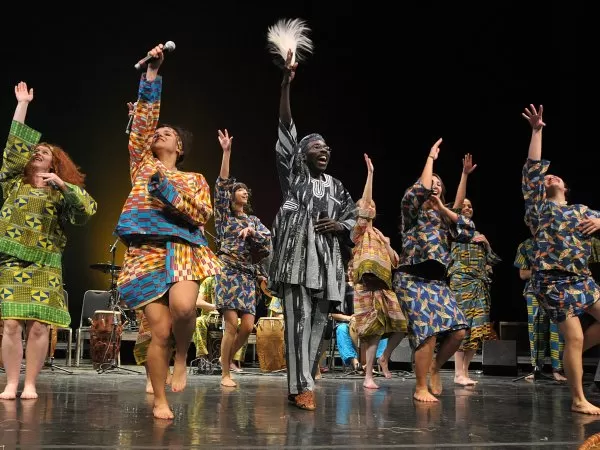 Performers in colorful West African-inspired attire sing and dance on stage during a lively musical performance.