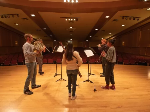 Five musicians rehearse on stage in an auditorium, standing in a semicircle with music stands and instruments.