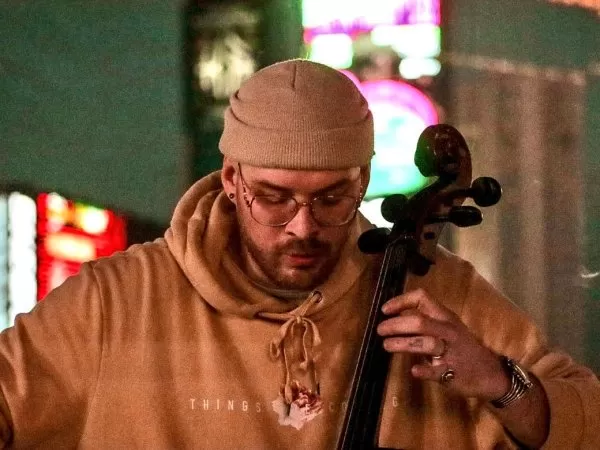 Cellist wearing a tan beanie, glasses and a brown hoodie plays the cello in a nighttime setting with colorful lights in the background.