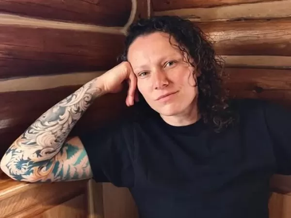 Person with curly hair and a tattooed arm, sitting against a wooden wall.