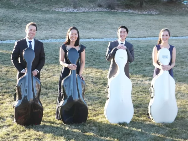 Four musicians stand outdoors on a grassy lawn, each holding a cello case.