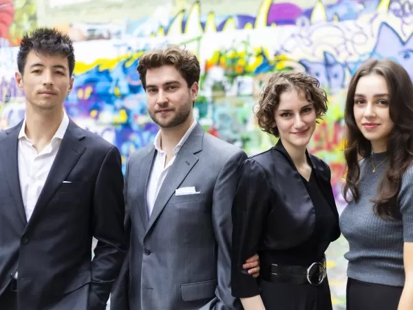 Four musicians pose for a group portrait in front of a colorful graffiti-covered wall, dressed in formal and semi-formal attire.