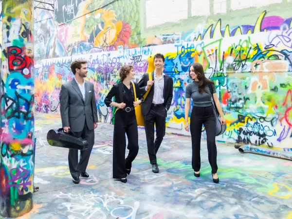 Four musicians walk through a colorful graffiti-covered alley, carrying instrument cases and bags.