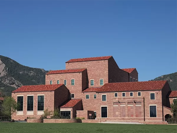 Exterior of the Imig Music Building on the CU Boulder campus, with the Flatirons rising behind it under a clear blue sky.